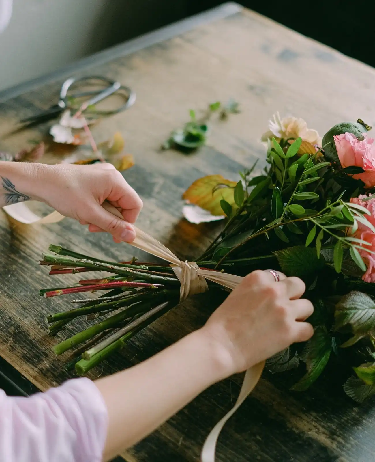A florist&rsquo;s hands tie a ribbon around the stems of a bridal bouquet on a wooden bench.